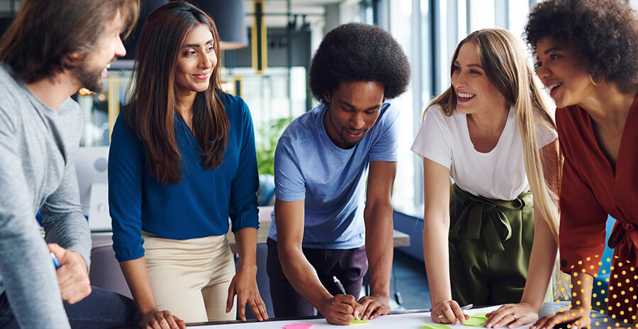 5 pessoas diferentes em pé em volta de uma mesa conversando e trabalhando e escrevendo em papéis