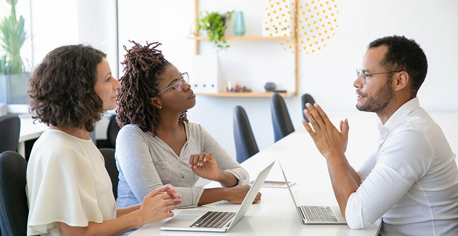 imagem de duas mulheres sentadas de frente para um homem com dois notebooks na mesa conversando