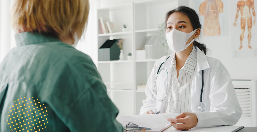 imagem de uma médica usando máscara sentada conversando com uma paciente e segurando um papel