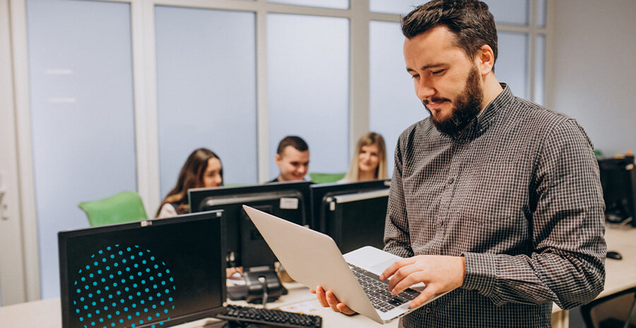 imagem de um homem em pé segurando um computador com três pessoas sentadas ao fundo