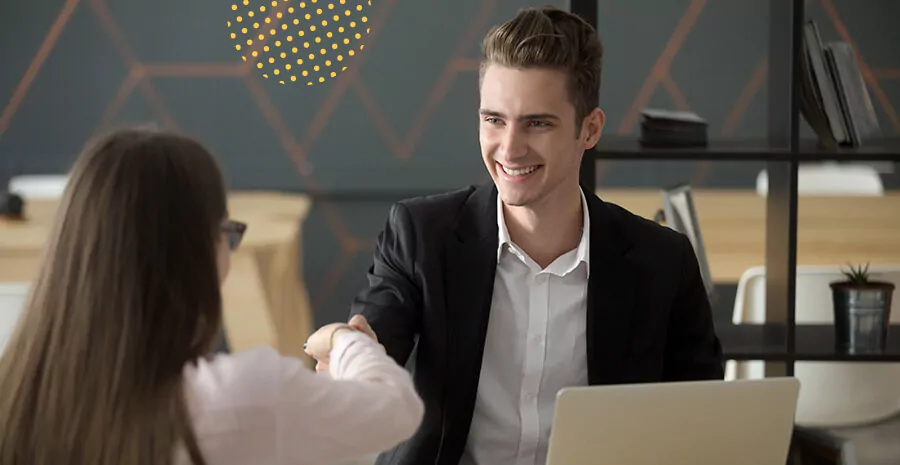 imagem de um homem sentado sorrindo em frente um notebook cumprimentando uma mulher com aperto de mãos