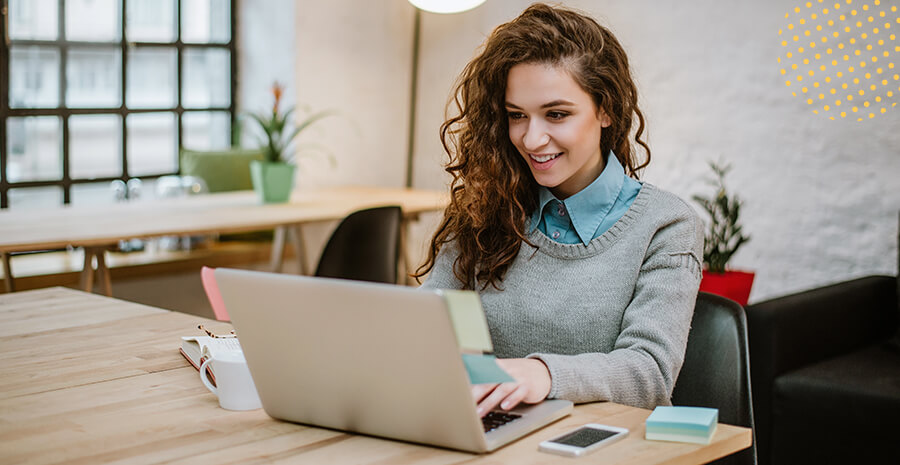 imagem de uma mulher sentada na frente de um computador sorrindo e com o celular do lado
