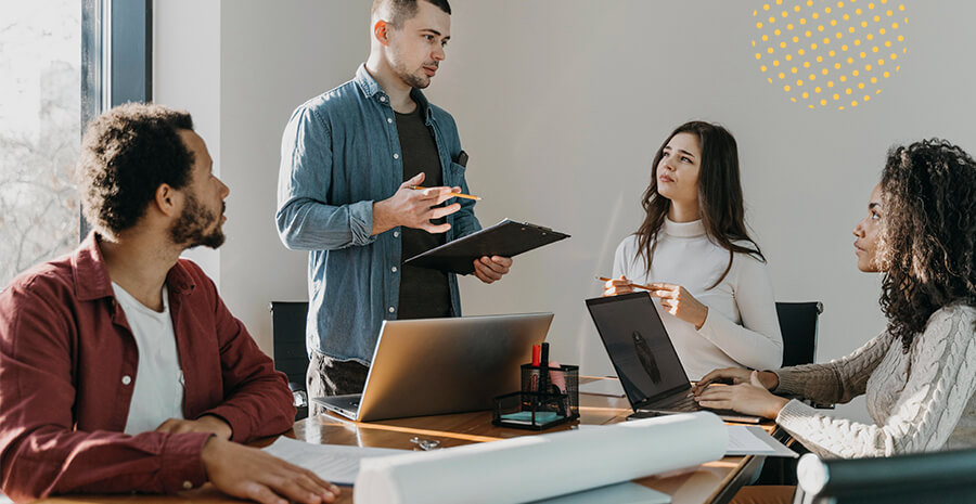 imagem de um homem em pé conversando com outro homem e duas mulheres sentadas ao redor de uma mesa na frente de computadores