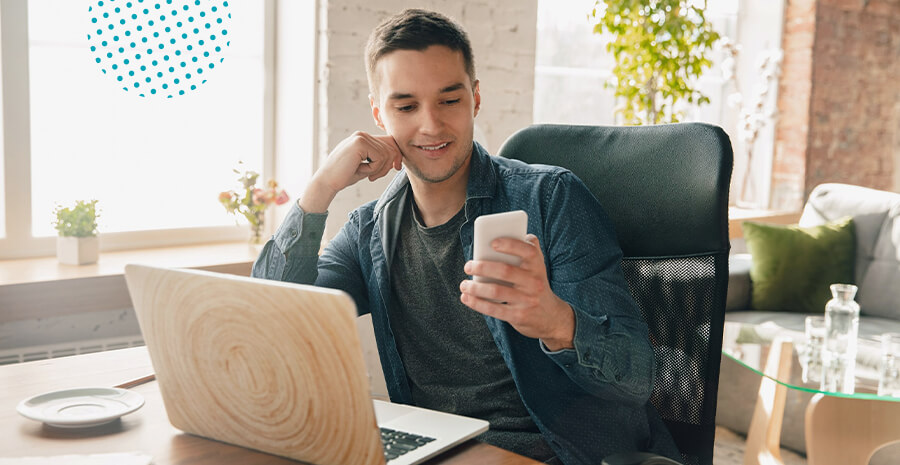 imagem de um homem sorrindo sentado em uma mesa de escritório segurando o celular e na frente do computador