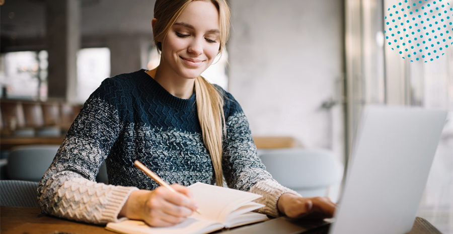 imagem de uma mulher sorrindo sentada na frente de um computador escrevendo em um caderno