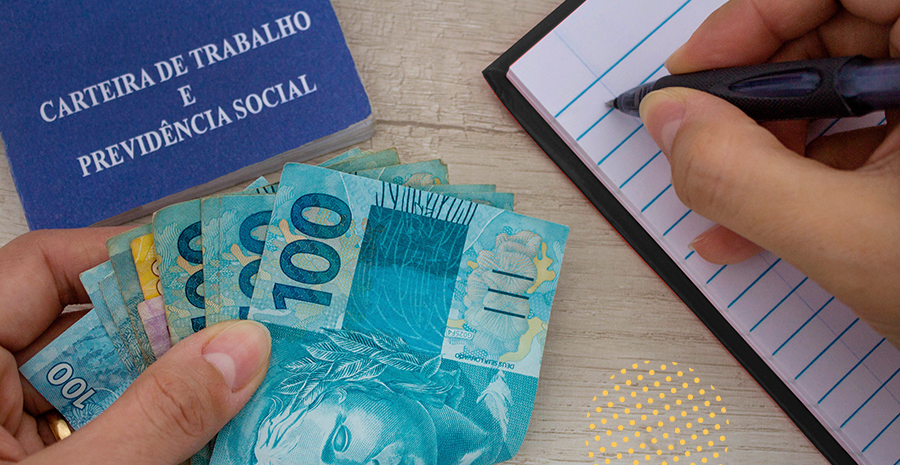 imagem de uma pessoa segurando notas de cem reais escrevendo em um caderno com a carteira de trabalho ao lado