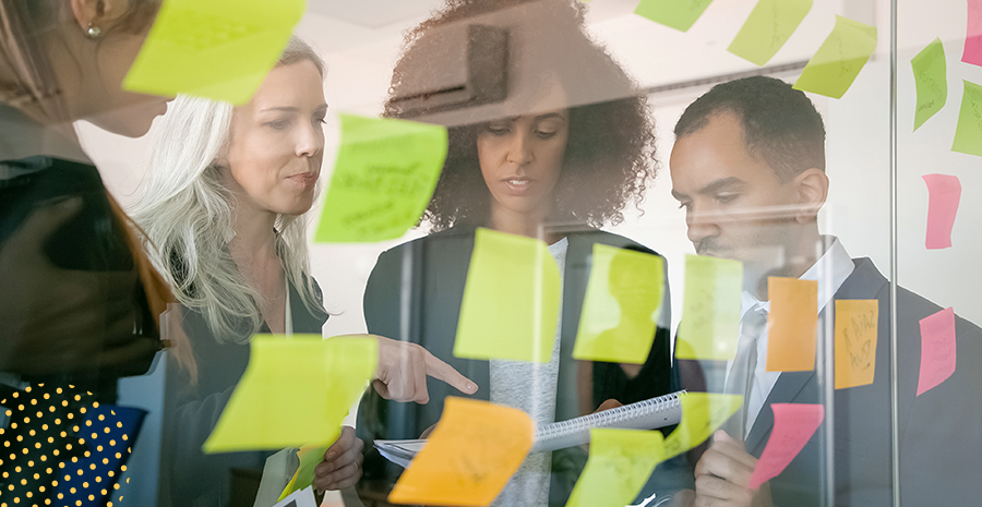 imagem de três mulheres e dois homens na frente uma parede de vidro com postits colados
