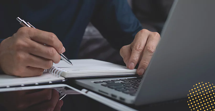 imagem de um homem sentado na frente de um computador escrevendo em um caderno
