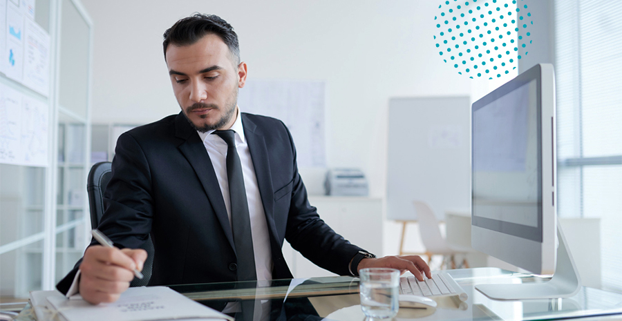 imagem de um homem sentado em uma mesa digitando em um computador e escrevendo em um caderno