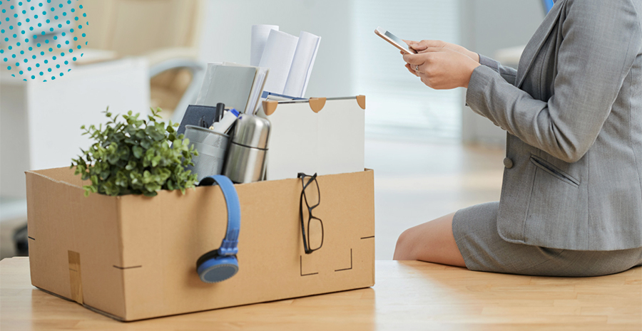 imagem de uma mulher sentada em cima de uma mesa ao lado de uma caixa de papelão com cadernos, um óculos e uma planta dentro