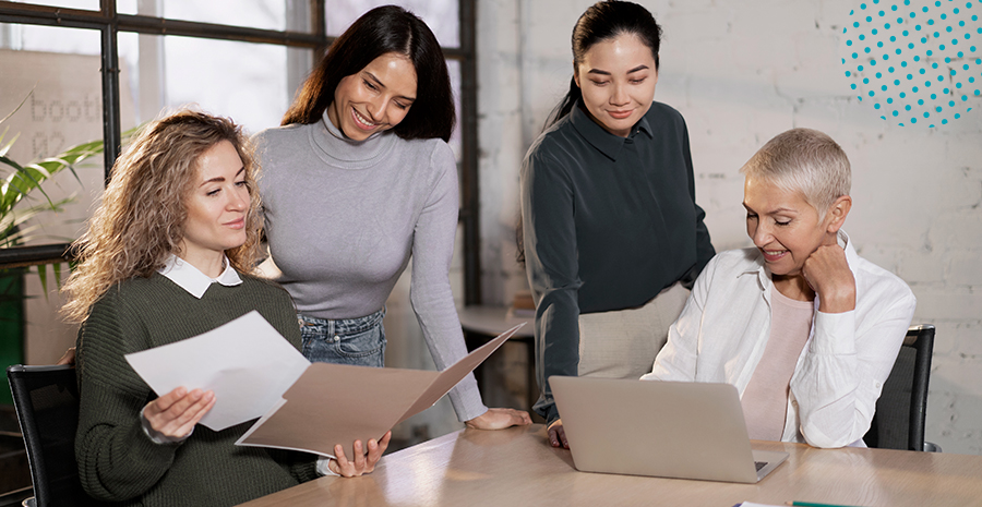 imagem de quatro mulheres ao redor de uma mesa conversando e olhando para papéis e um computador