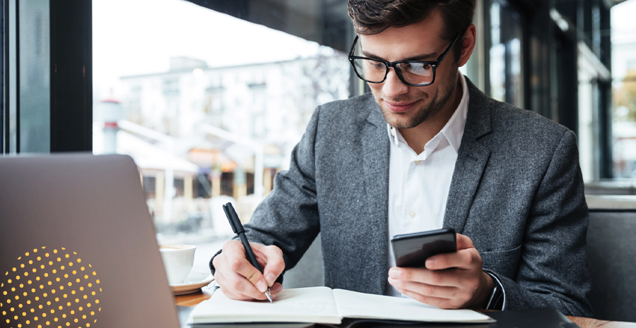 imagem de um homem sentado na frente de um computador segurando um celular e escrevendo em um caderno