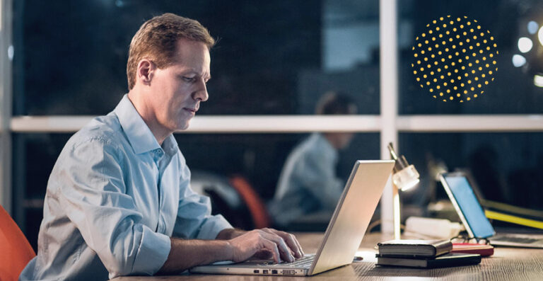 imagem de um homem teclado em um notebook sob uma mesa, com luminária acesa realizando hora extra noturna