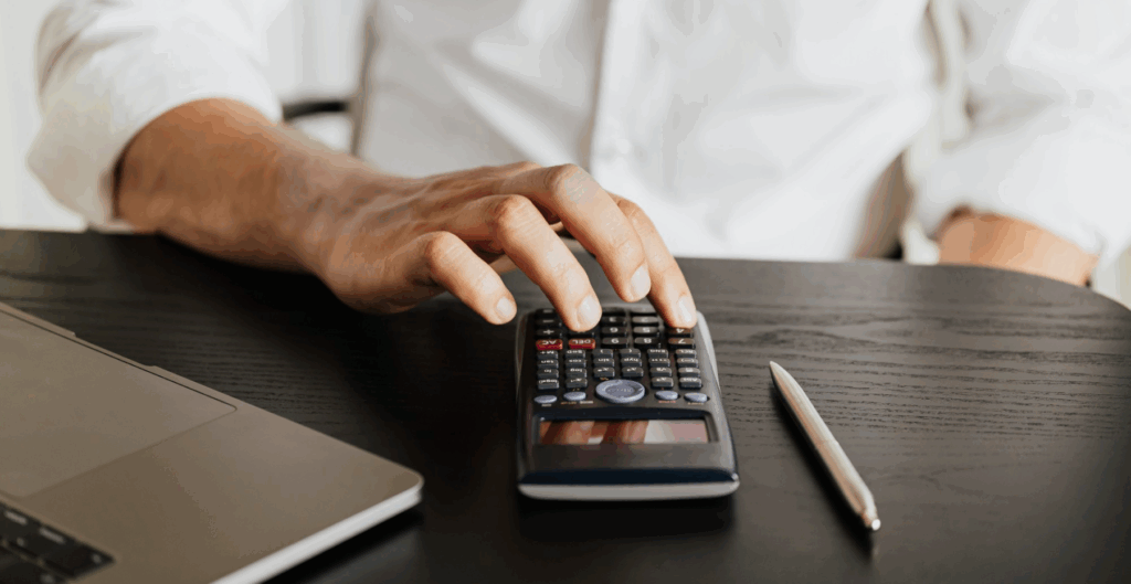 Pessoa usando calculadora em uma mesa de escritório, com laptop e caneta ao lado, ambiente de escritório, foco em tecnologia e gadgets.