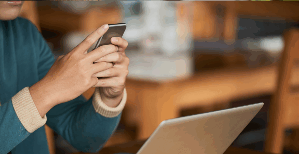 Pessoa utilizando smartphone em ambiente de trabalho com laptop aberto ao lado, foco no aparelho móvel, fundo de escritório com móveis de madeira.