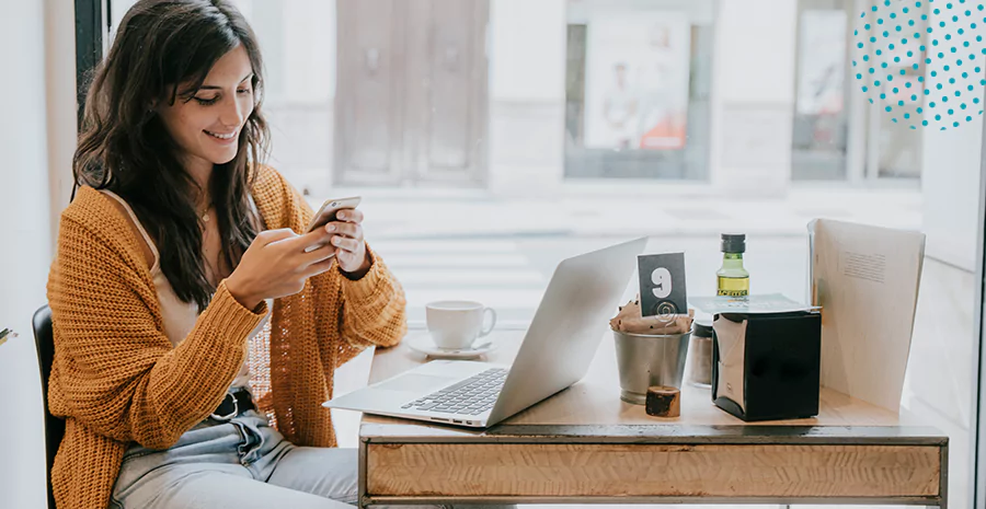 mulher segurando seu celular e sorrindo em frente a uma mesa que tem um notebook, xicara e café e outras utensílios de escritório